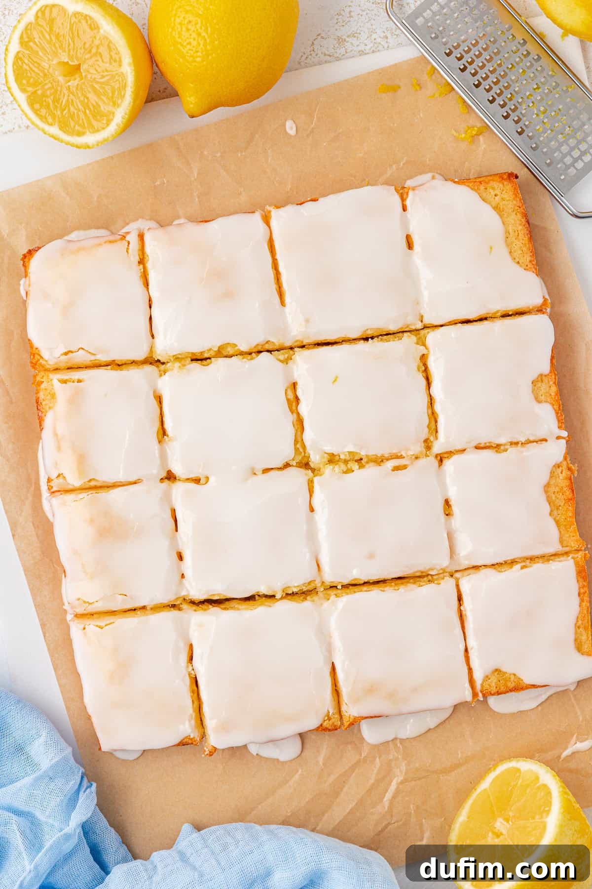 Zesty Lemon Brownies 14 A batch of lemon brownies with white icing is cut into squares on parchment paper. Whole and halved lemons, a zester, and a light blue cloth are nearby on the white surface.