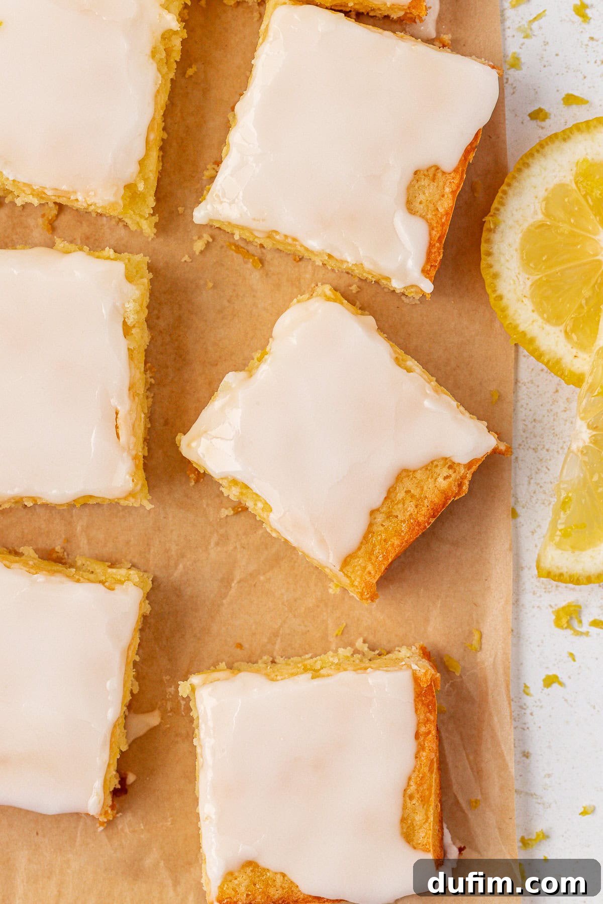 Zesty Lemon Brownies 15 Several lemon brownies with glossy white icing are arranged on brown parchment paper, with a couple of fresh lemon slices nearby. The bars have a light, golden color and look soft and moist.