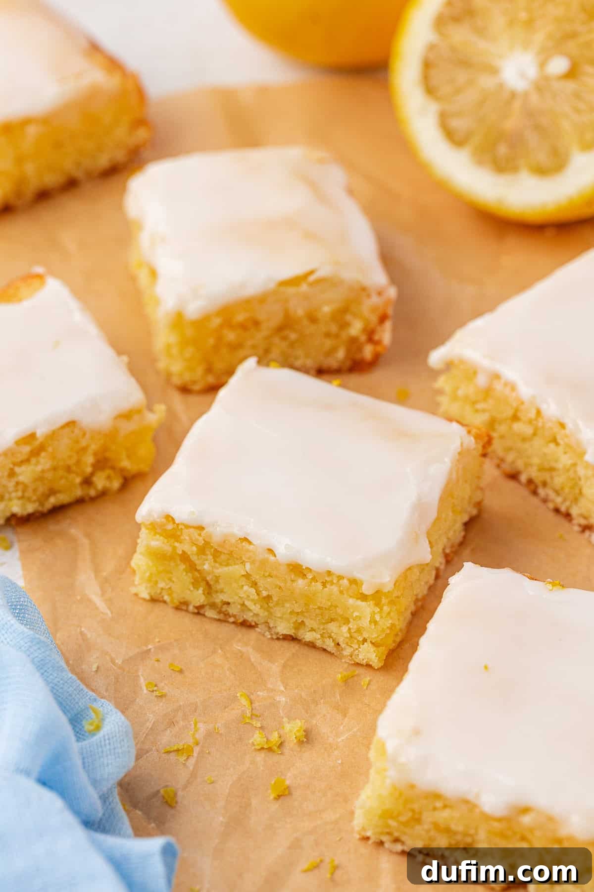 Zesty Lemon Brownies 3 Six square lemon brownies with glossy white icing are arranged on brown parchment paper. A vibrant blue napkin and a halved lemon add a fresh, inviting touch to the scene.