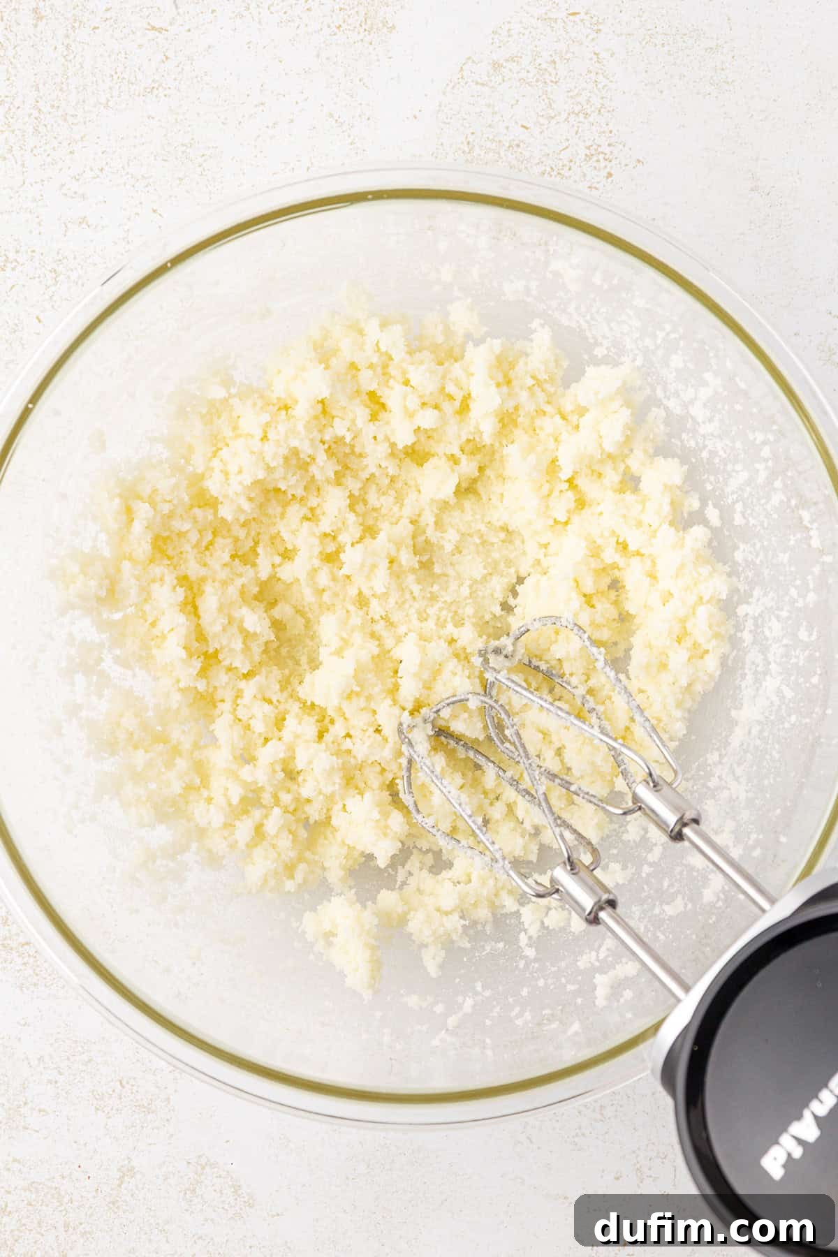 Zesty Lemon Brownies 6 A glass bowl with creamed butter and sugar being mixed by a handheld electric mixer on a light, textured surface.