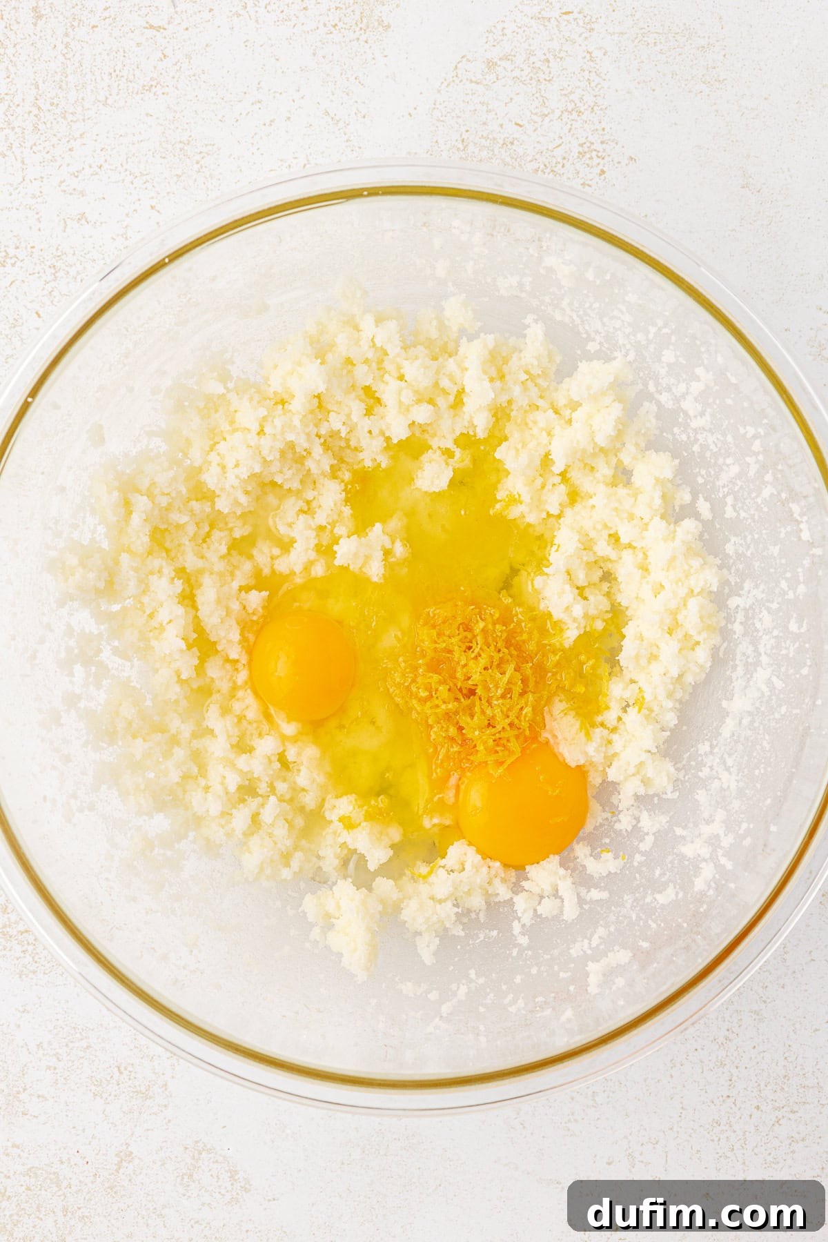 Zesty Lemon Brownies 7 A glass bowl containing creamed butter and sugar, two raw eggs, and grated lemon zest, set on a white surface.