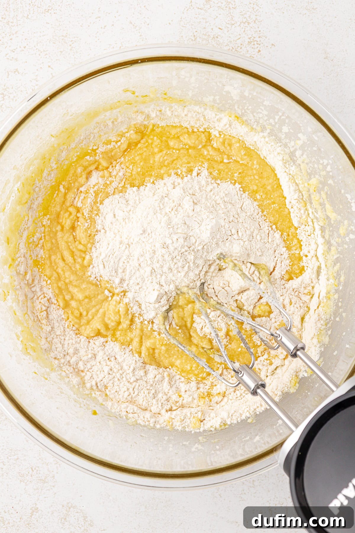 Zesty Lemon Brownies 8 A close-up of a mixing bowl containing cake batter with a pile of flour on top, and an electric hand mixer with beaters partially submerged in the mixture.