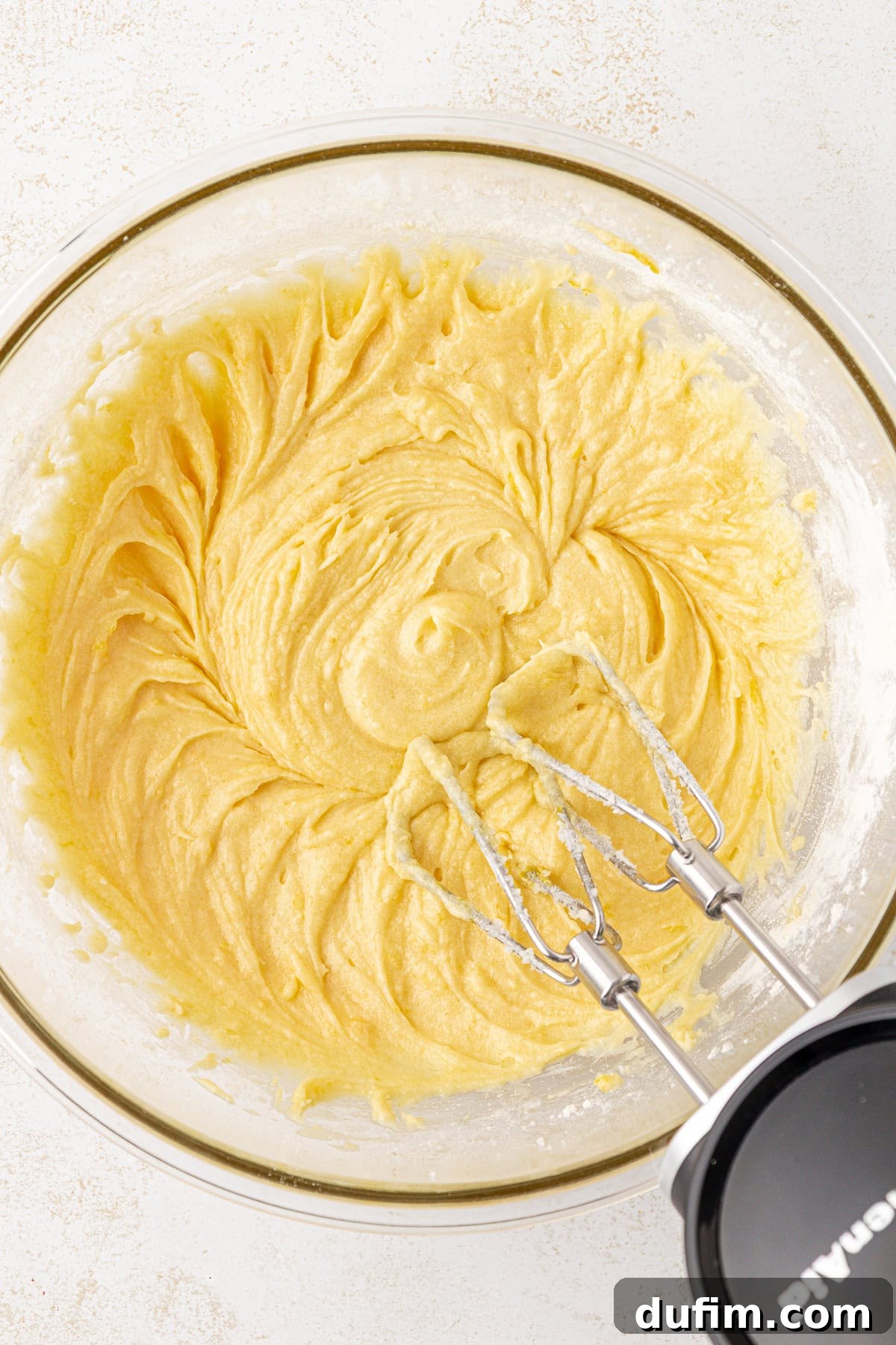 Zesty Lemon Brownies 9 A glass bowl filled with creamy yellow cake batter being mixed with an electric hand mixer; the beaters are resting in the batter.