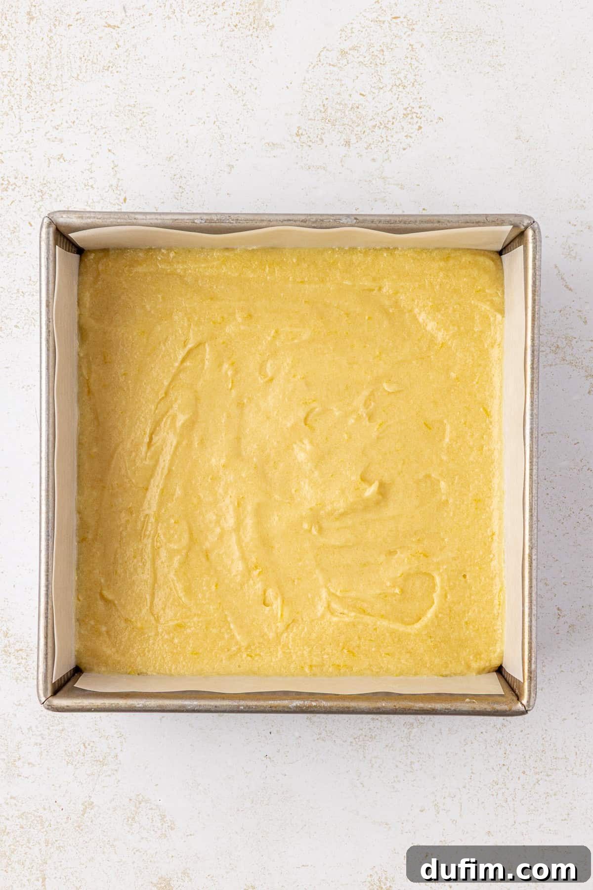 Zesty Lemon Brownies 10 A square baking pan lined with parchment paper is filled with an even layer of smooth, yellow cake batter, ready to be baked.