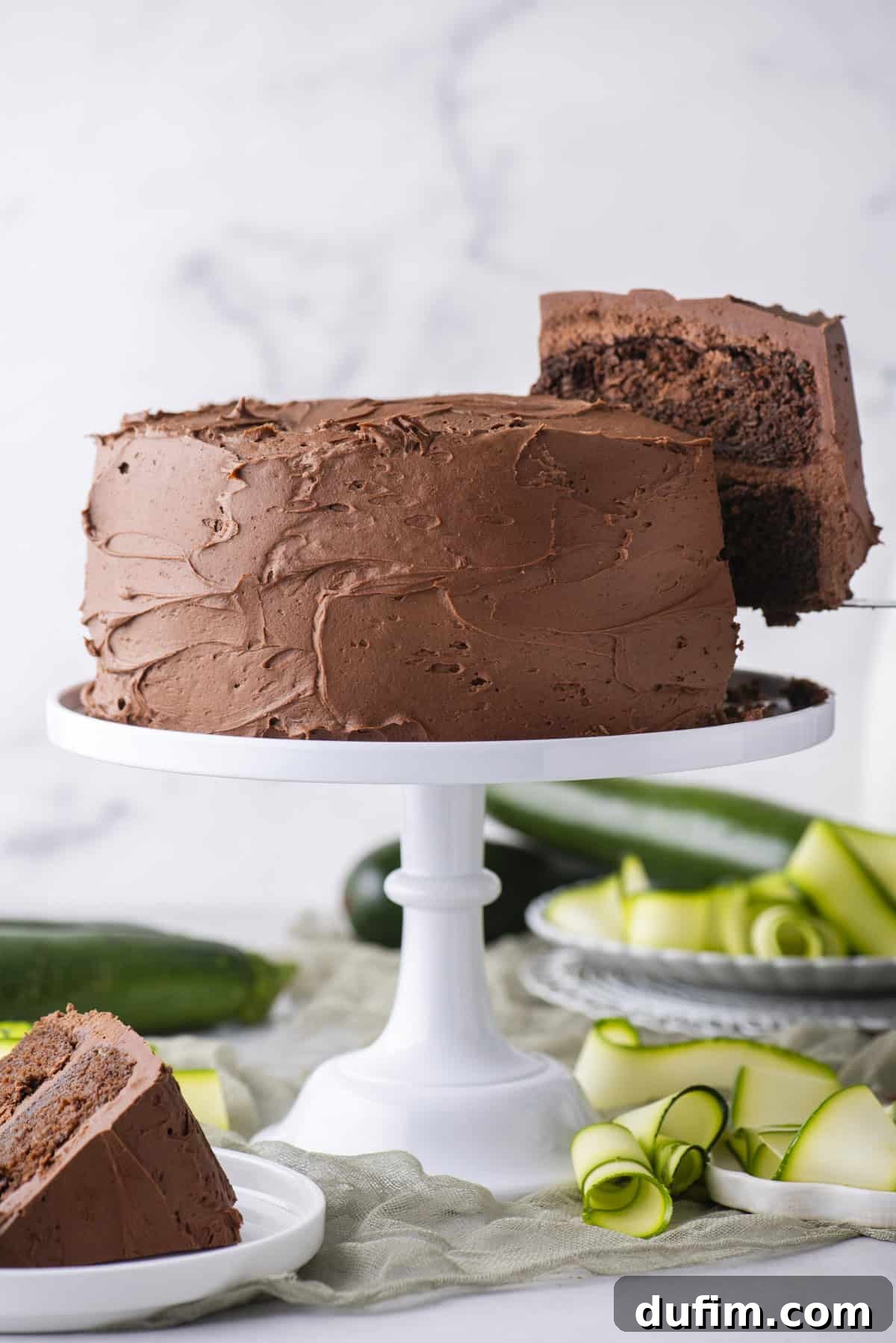 A chocolate frosted layer cake sits on a white cake stand, with one slice being served. Zucchini ribbons and a plate with another cake slice are nearby on a light-colored surface.