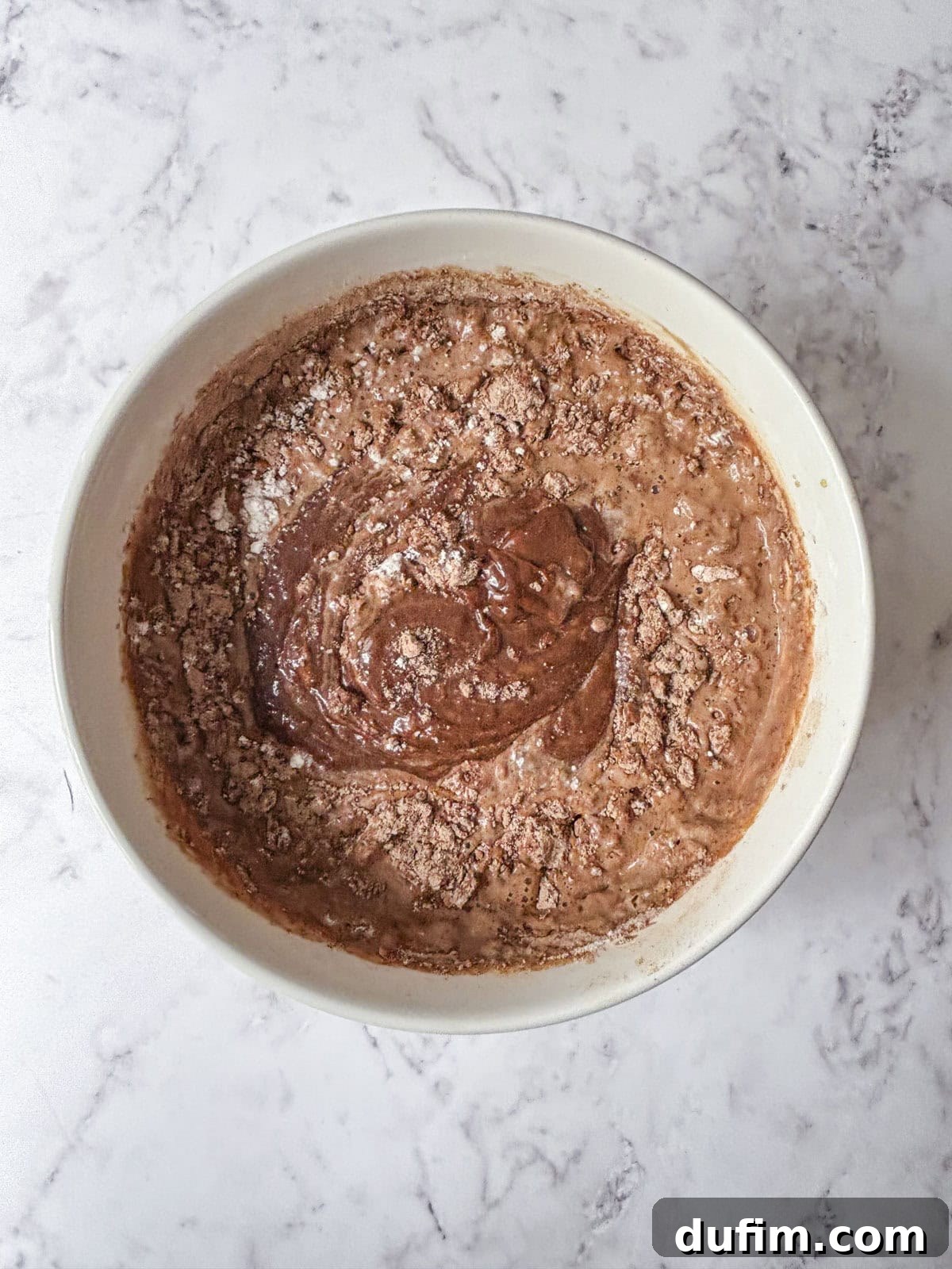 A white bowl filled with partially mixed chocolate cake batter sits on a light marble surface. Dry cocoa powder and flour are still visible on top of the wet ingredients.