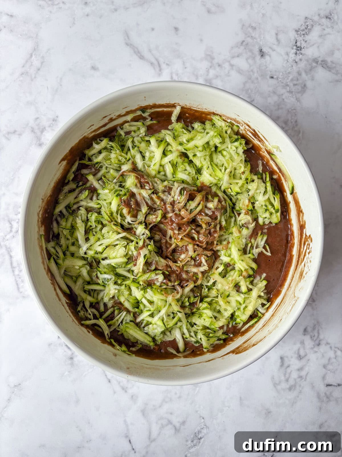 Overhead view of a mixing bowl with shredded zucchini being mixed into chocolate cake batter on a light marble countertop.