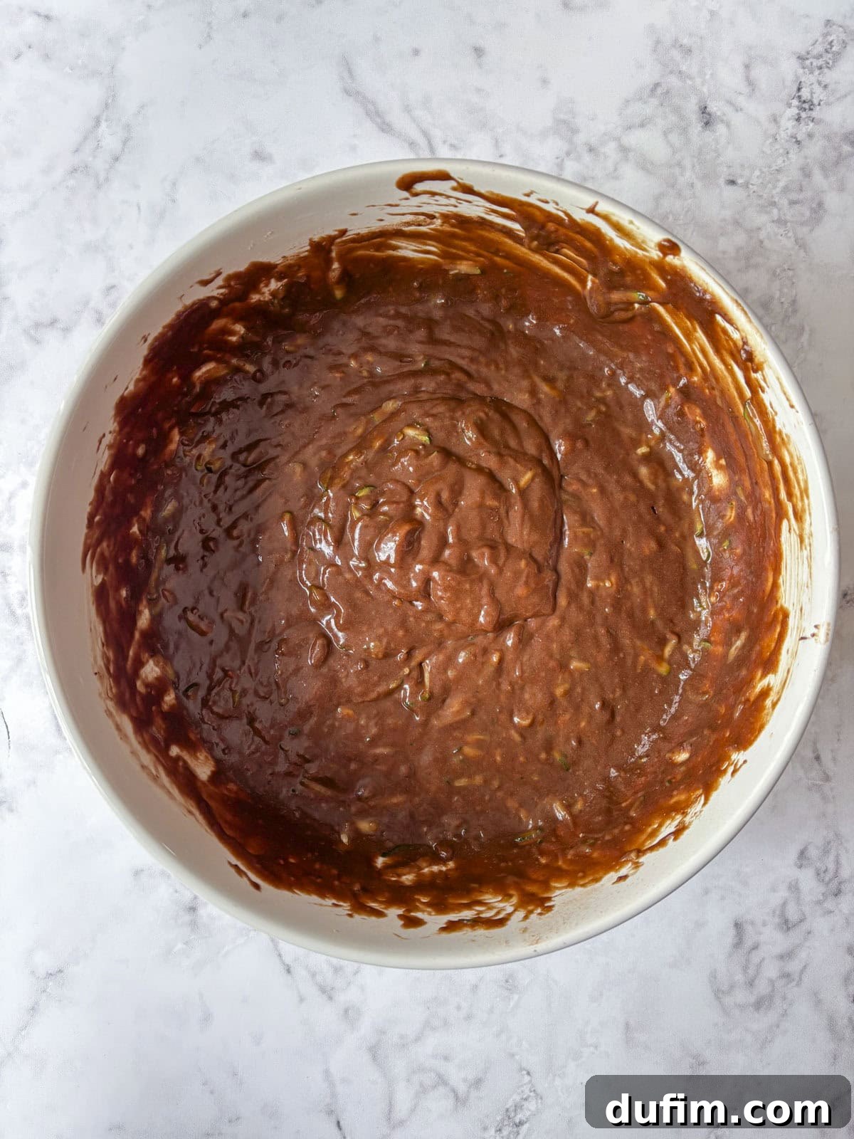 A white bowl filled with chocolate cake or brownie batter sits on a light marble countertop. The batter appears thick and slightly textured, ready to be baked.