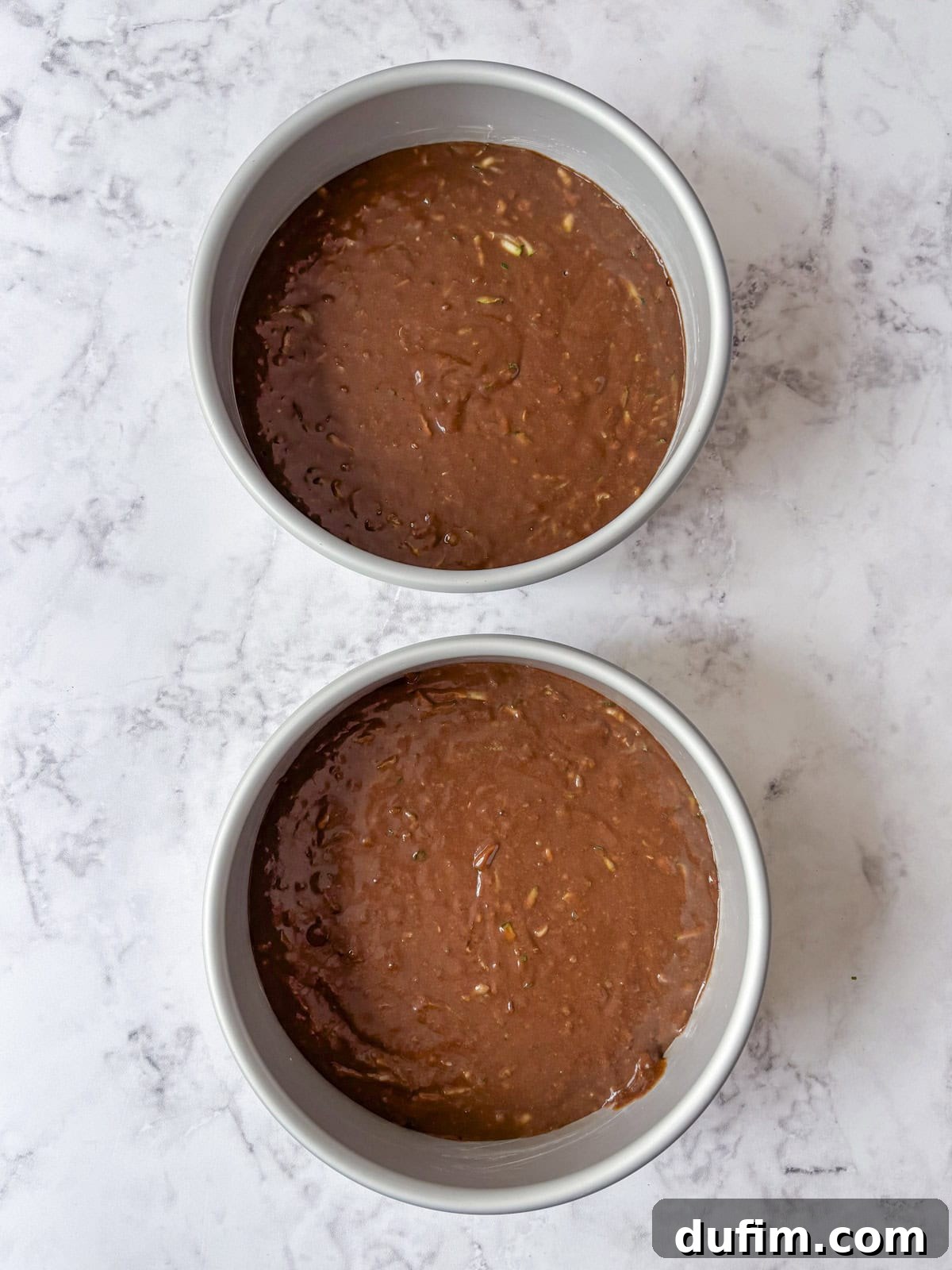 Two round cake pans filled with chocolate cake batter sit on a white marble surface, ready to be baked.