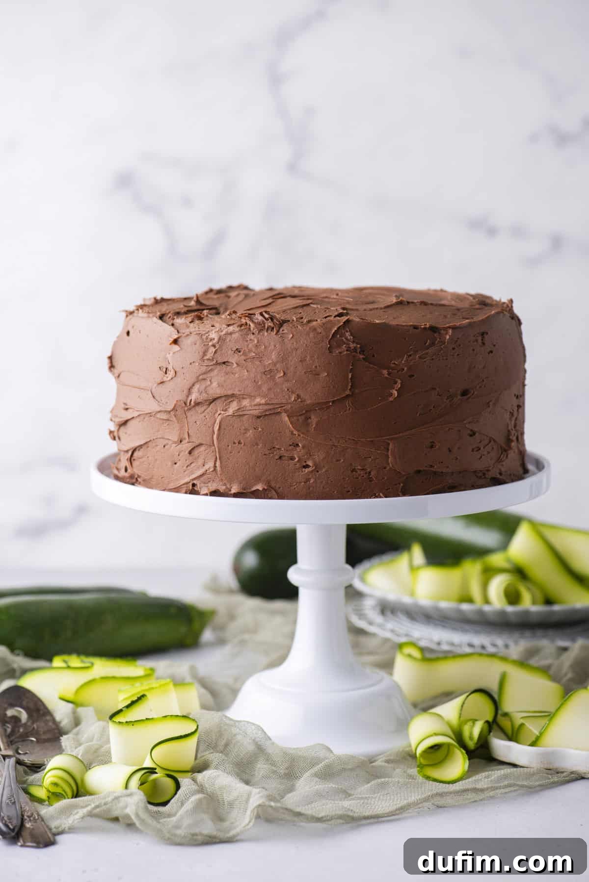 A chocolate frosted layer cake sits on a white cake stand, surrounded by green zucchini ribbons and whole zucchinis on a light surface with a marble background.