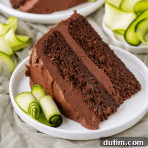 A slice of chocolate cake with chocolate frosting is served on a white plate, garnished with thin, rolled slices of zucchini. More zucchini rolls and a second plate are visible in the background.