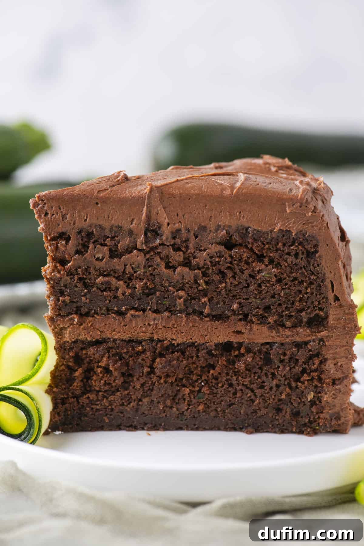 A close-up of a slice of chocolate layer cake with chocolate frosting on a white plate, garnished with a curl of yellow and green zucchini. The background is softly blurred.