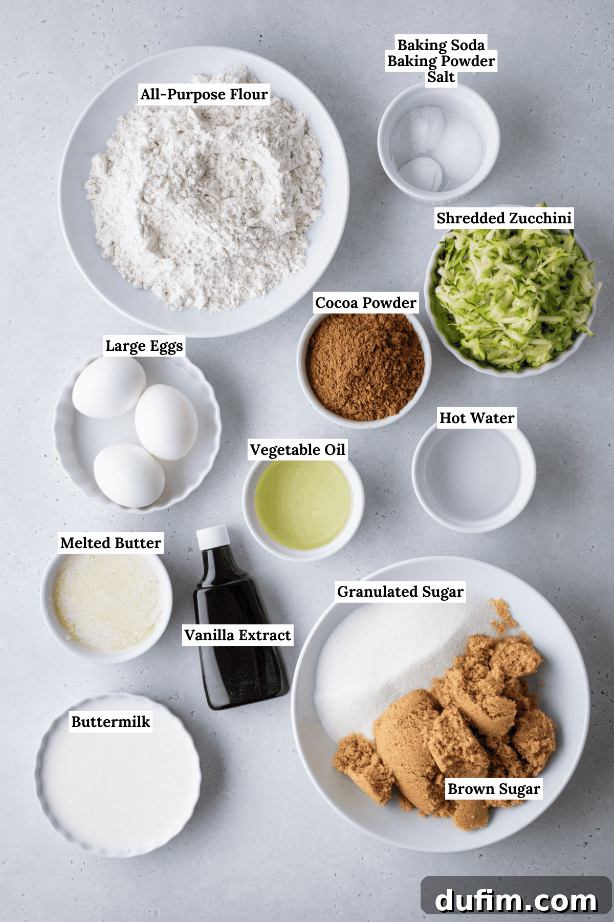 Top-down view of labeled baking ingredients in bowls on a gray surface, including flour, eggs, buttermilk, melted butter, oil, vanilla, granulated and brown sugars, cocoa powder, shredded zucchini, and a small bowl of leaveners.