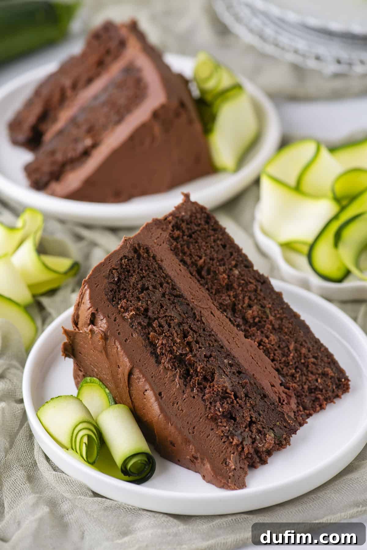 Two slices of rich chocolate cake with chocolate frosting are served on white plates, each garnished with thinly sliced, rolled zucchini ribbons. A stack of plates is blurred in the background.