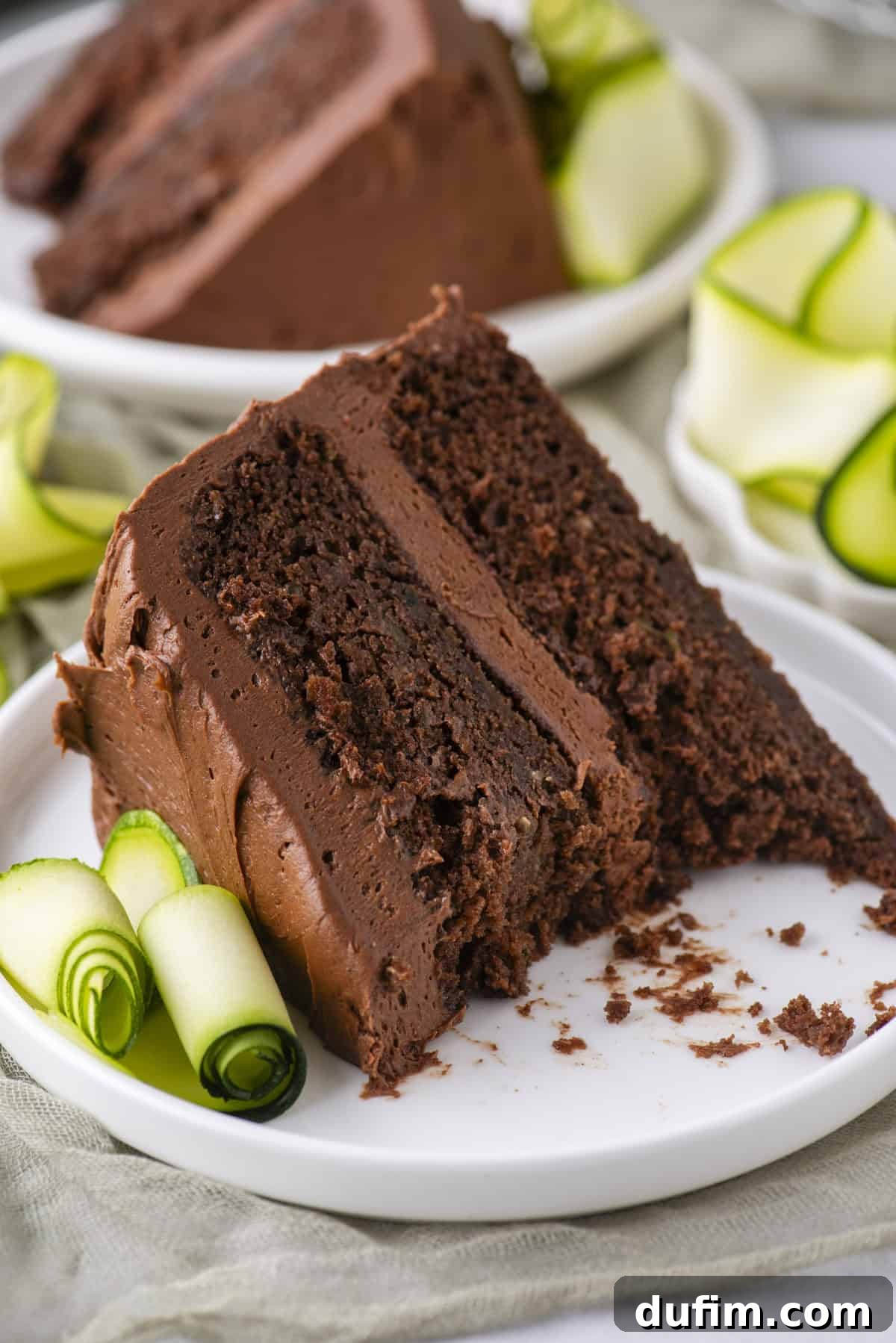 A slice of rich chocolate cake with chocolate frosting on a white plate, garnished with rolled slices of zucchini, with another plate of cake in the background.