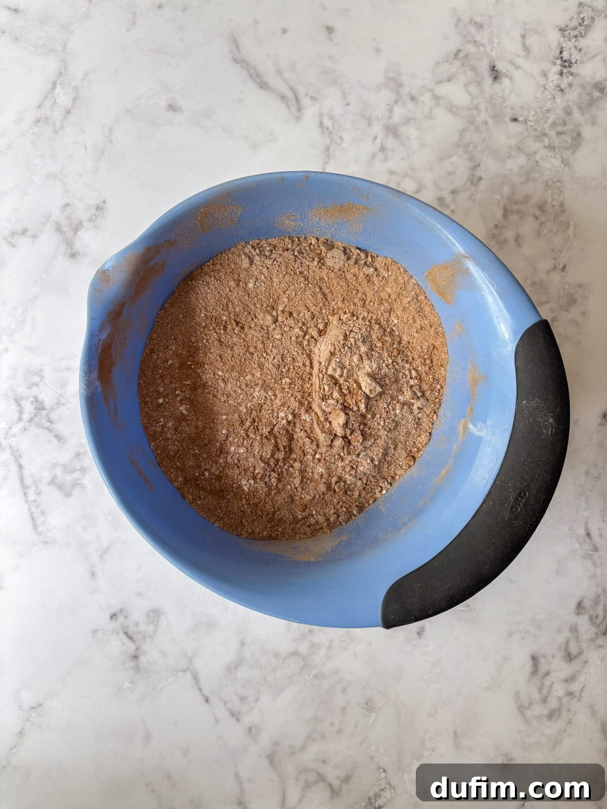 A blue mixing bowl with a black handle contains a blended mixture of dry cocoa powder and flour on a white marble surface.
