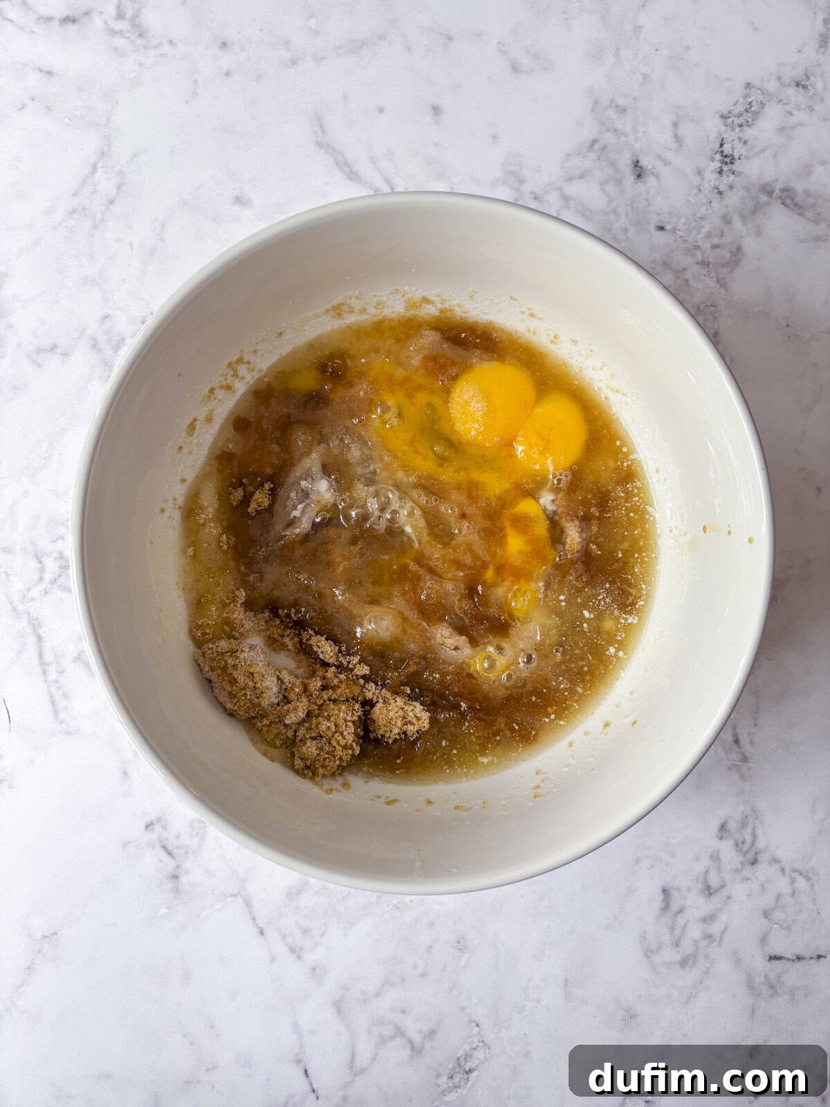 A white bowl on a marble surface containing eggs, brown sugar, and wet ingredients about to be mixed together.