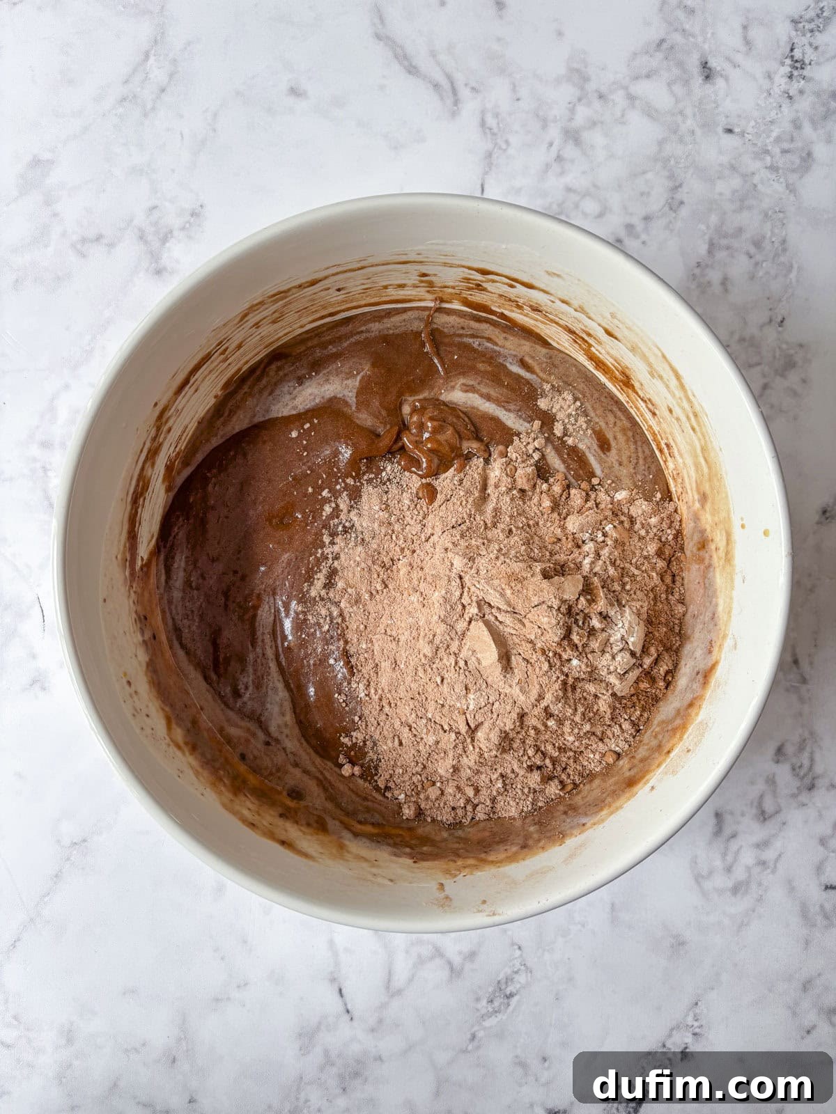 A white mixing bowl on a marble surface contains chocolate batter, with some dry cocoa powder still visible on top, not yet fully mixed in.