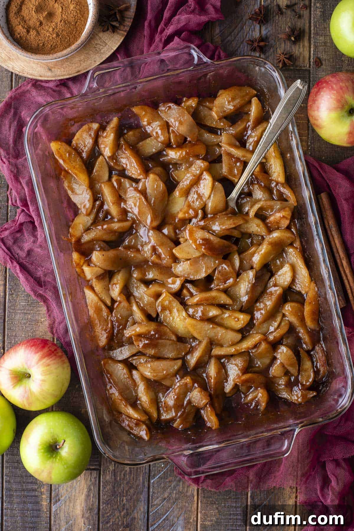 Warm Spiced Baked Apples 3 A glass baking dish filled with baked cinnamon apple slices sits on a wooden table, surrounded by fresh apples, cinnamon sticks, a maroon cloth, and a bowl of ground cinnamon. A serving spoon rests in the dish.