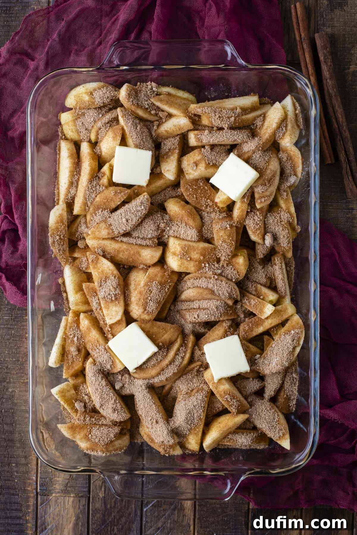 Warm Spiced Baked Apples 5 A glass baking dish filled with sliced apples topped with a layer of cinnamon sugar and four cubes of butter, set on a rustic wooden surface with a deep red cloth and cinnamon sticks beside it.