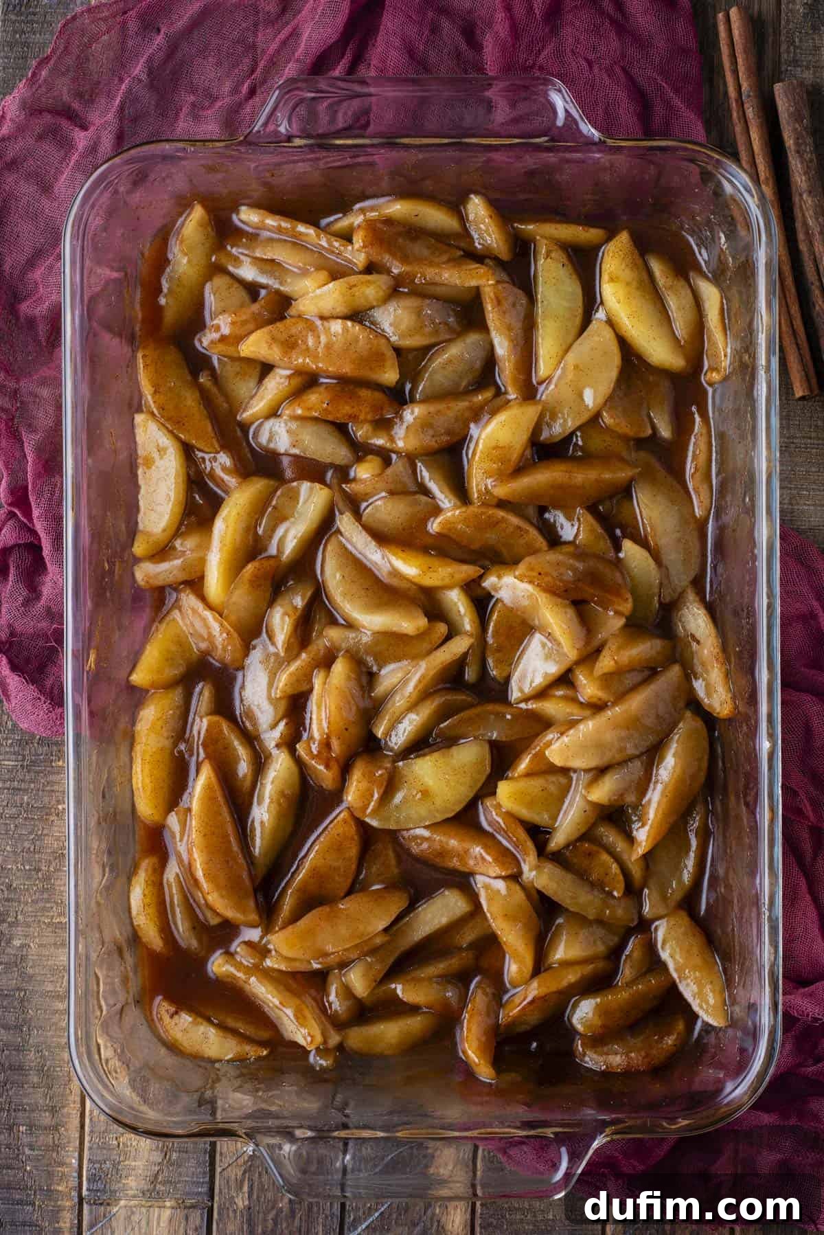Warm Spiced Baked Apples 6 A glass baking dish filled with sliced apples coated in a glossy cinnamon sauce, sitting on a wooden surface with a dark red cloth and cinnamon sticks beside it.