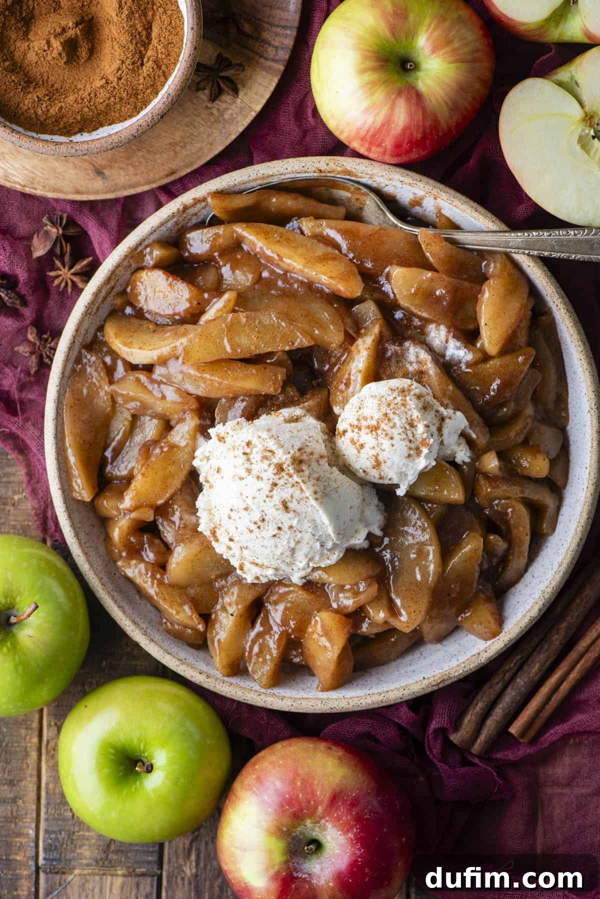 Warm Spiced Baked Apples 7 A bowl of baked cinnamon apples topped with two scoops of vanilla ice cream and sprinkled with cinnamon, surrounded by fresh apples, cinnamon sticks, and a bowl of ground cinnamon.