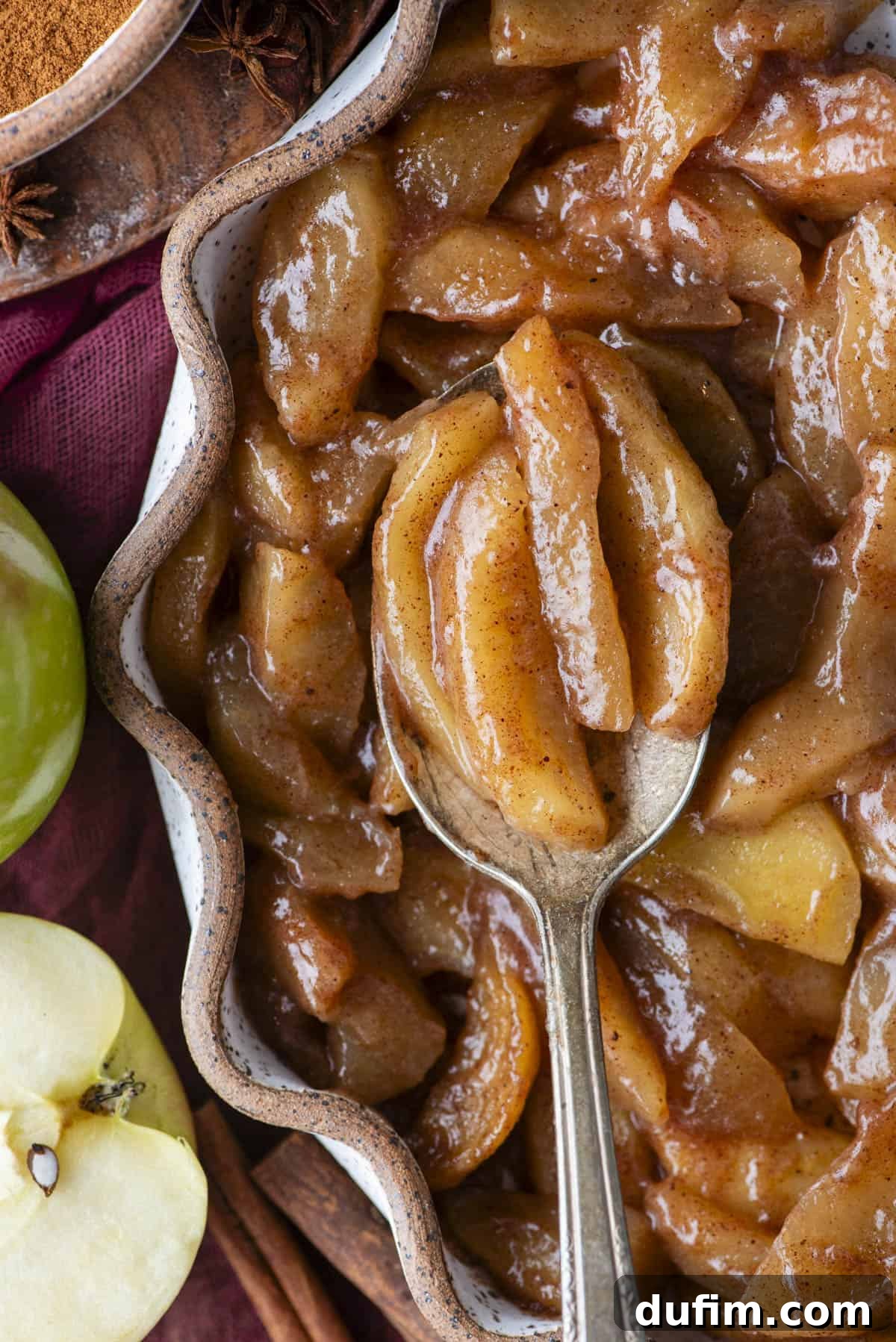 Warm Spiced Baked Apples 8 A close-up of baked cinnamon apples in a wavy-edged dish, with a spoon lifting several glazed apple slices. Green apples and cinnamon sticks are nearby.