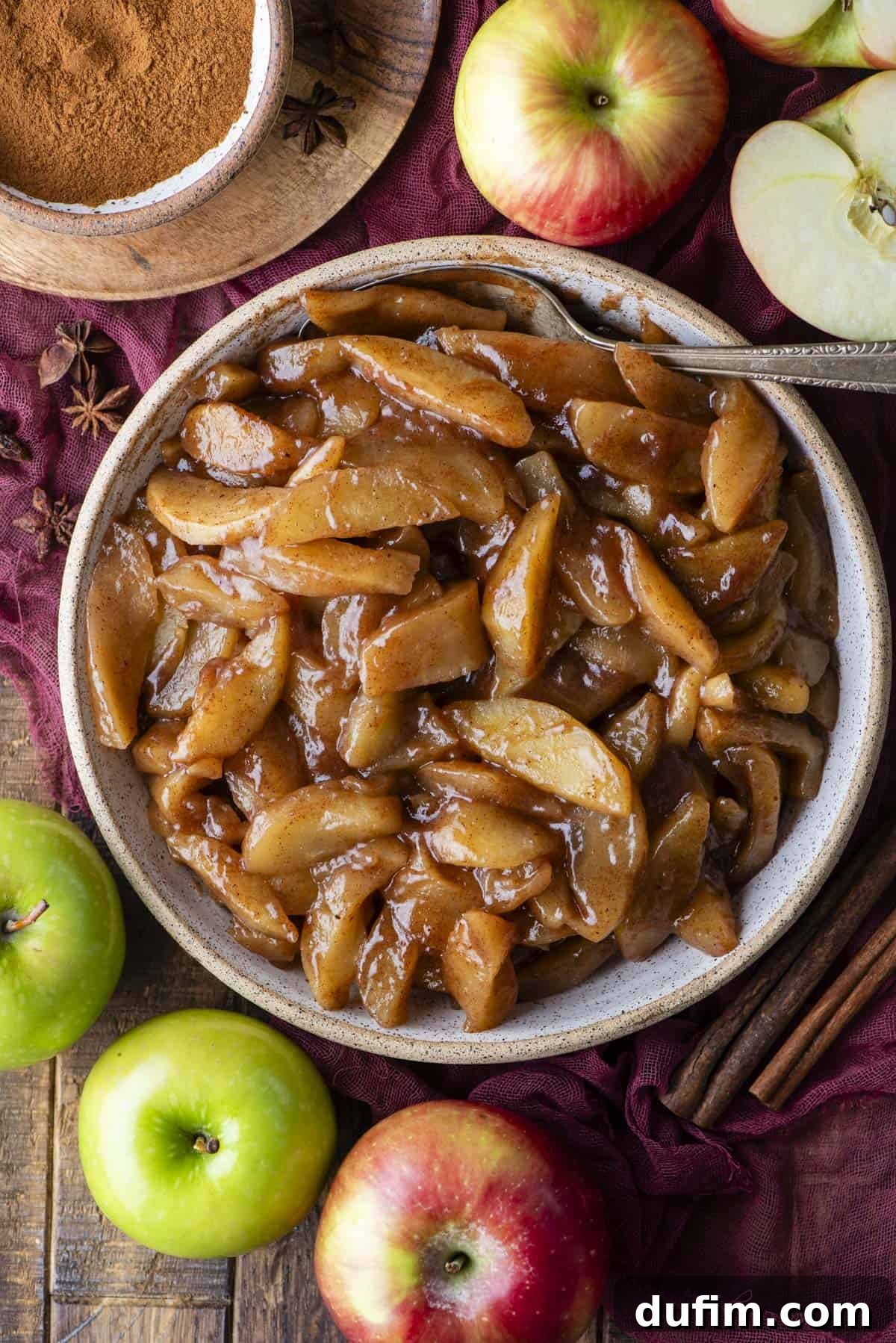Warm Spiced Baked Apples 9 A bowl of cooked cinnamon apples with a spoon, surrounded by fresh red and green apples, cinnamon sticks, and a bowl of ground cinnamon on a rustic wooden surface.
