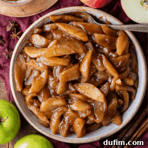Warm Spiced Baked Apples 10 A bowl filled with baked apples coated in a glossy brown cinnamon sauce sits on a rustic surface, surrounded by green apples, cinnamon sticks, and star anise. A silver spoon rests in the bowl.