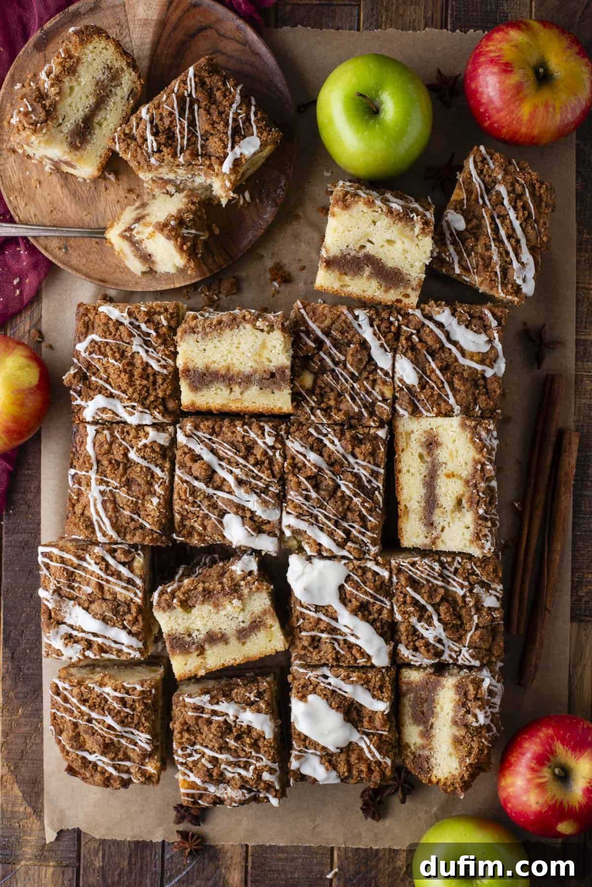 Apple Cinnamon Streusel Coffee Cake 2 Overhead view of sliced apple coffee cake squares with crumb topping and white icing drizzle, arranged on parchment paper. Green and red apples, cinnamon sticks, and star anise are scattered around for decoration.