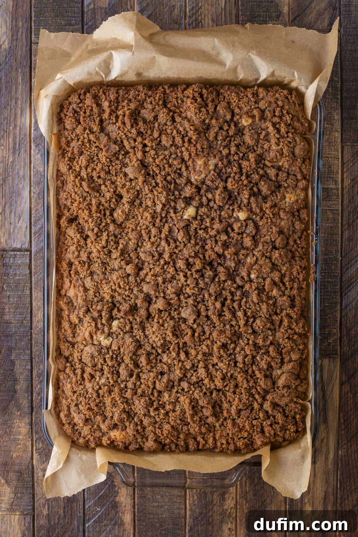 Apple Cinnamon Streusel Coffee Cake 8 A rectangular glass baking dish lined with parchment paper holds a golden-brown crumb cake topped with a thick, crumbly cinnamon streusel. The dish sits on a rustic wooden table.