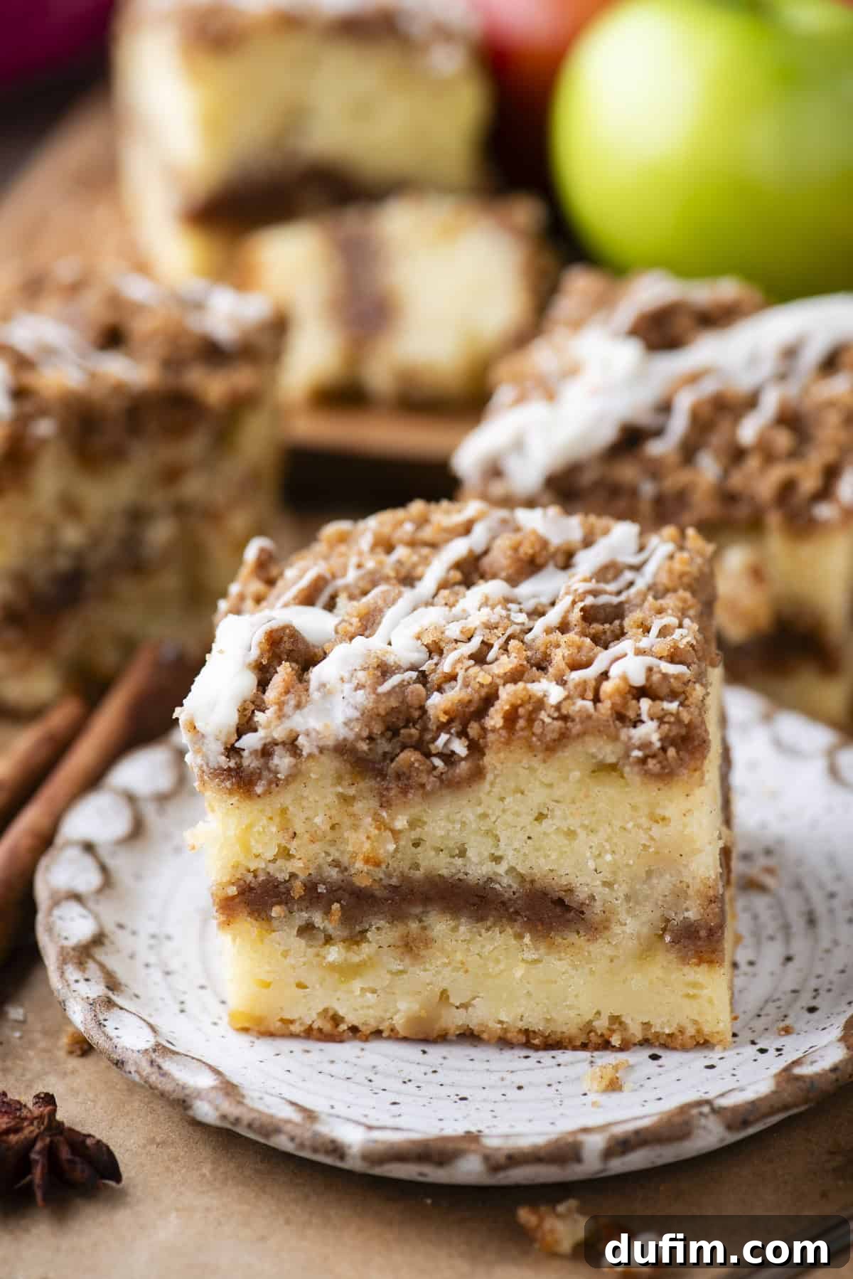 Apple Cinnamon Streusel Coffee Cake 10 A slice of crumbly coffee cake with a cinnamon swirl and a drizzle of white icing sits on a decorative plate, with more cake pieces and green apples in the background.