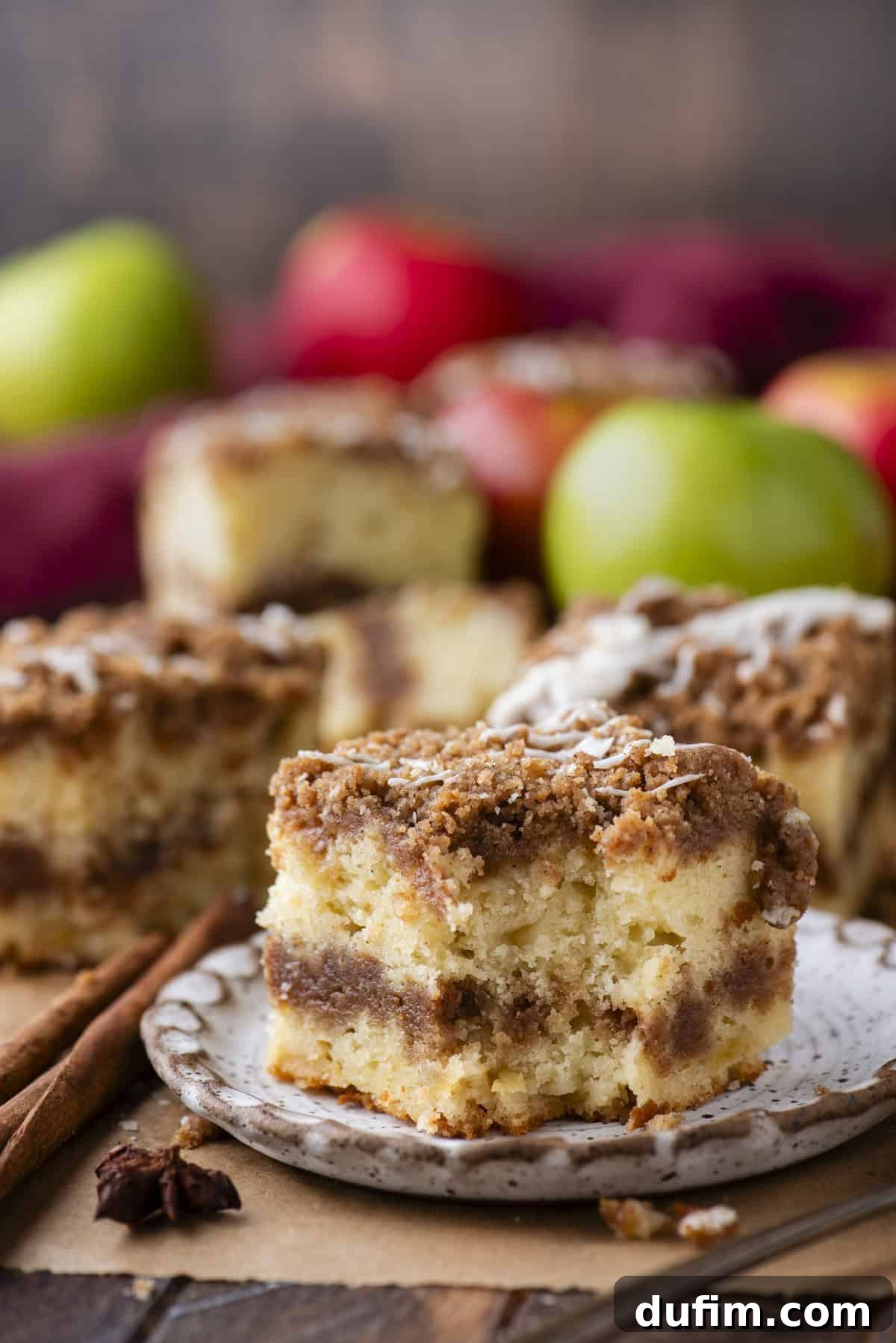 Apple Cinnamon Streusel Coffee Cake 11 A slice of crumbly coffee cake with cinnamon swirl on a plate, with more cake pieces and colorful apples blurred in the background.