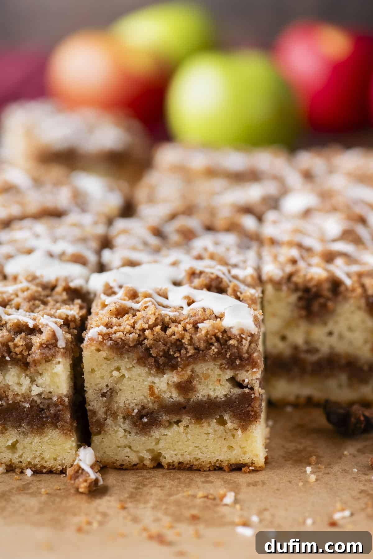 Apple Cinnamon Streusel Coffee Cake 4 Close-up of sliced coffee cake with a crumbly cinnamon streusel topping and white icing drizzle, with blurred apples in the background.