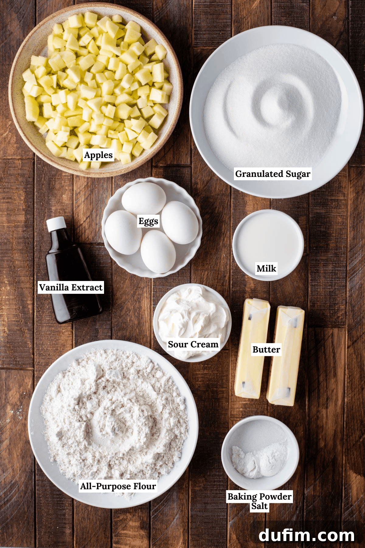 Apple Cinnamon Streusel Coffee Cake 6 Top-down view of ingredients for baking: cubed apples, granulated sugar, eggs, milk, vanilla extract, sour cream, two sticks of butter, all-purpose flour, baking powder, and salt on a wooden surface, each labeled.