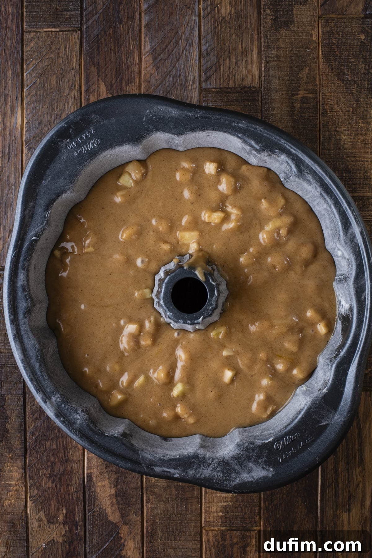 A bundt cake pan filled with light brown batter containing small apple chunks, sitting on a dark wooden surface. The pan is floured, and the batter is ready to be baked.