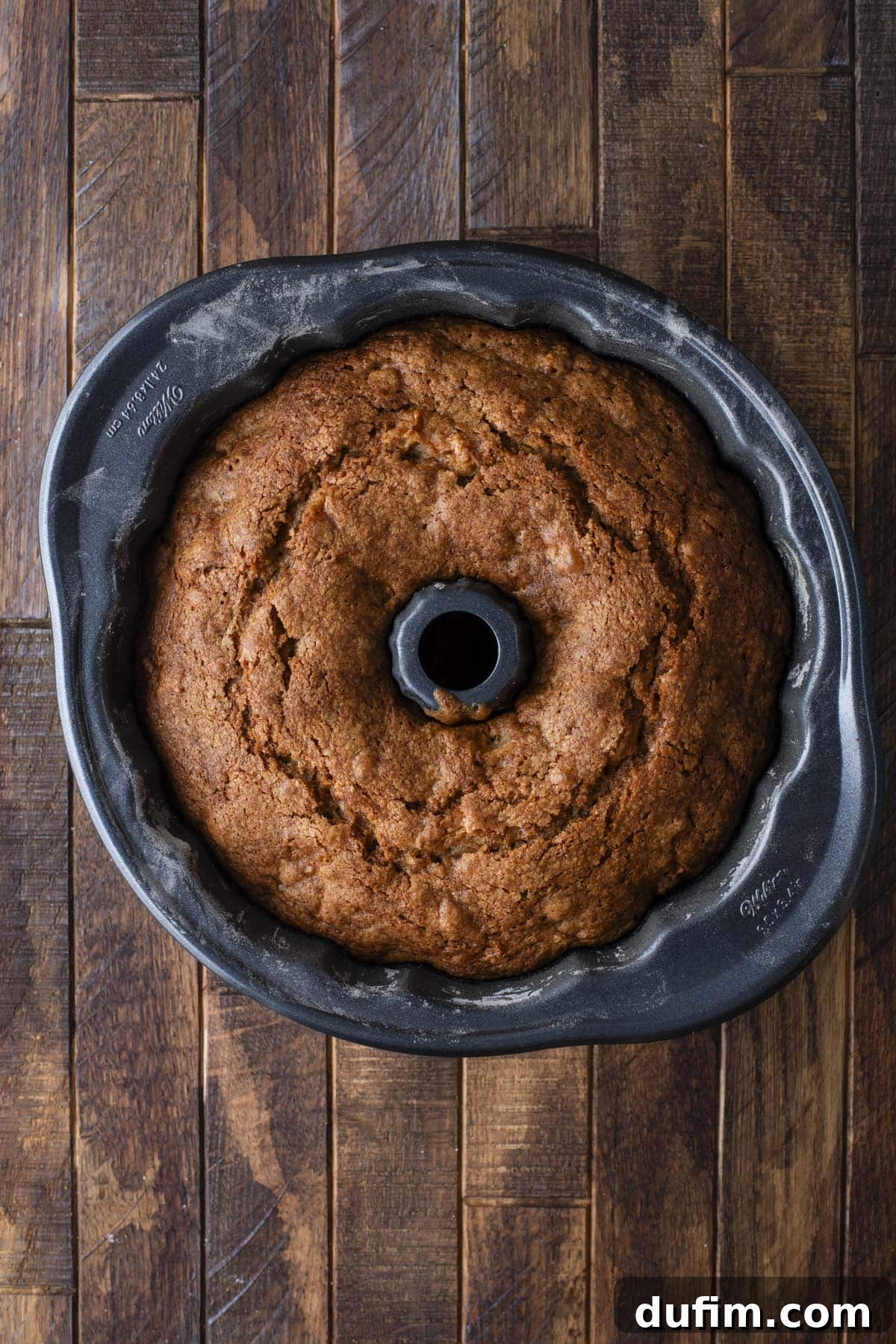 A golden-brown bundt cake sits freshly baked in a dark bundt pan on a wooden surface, viewed from above.