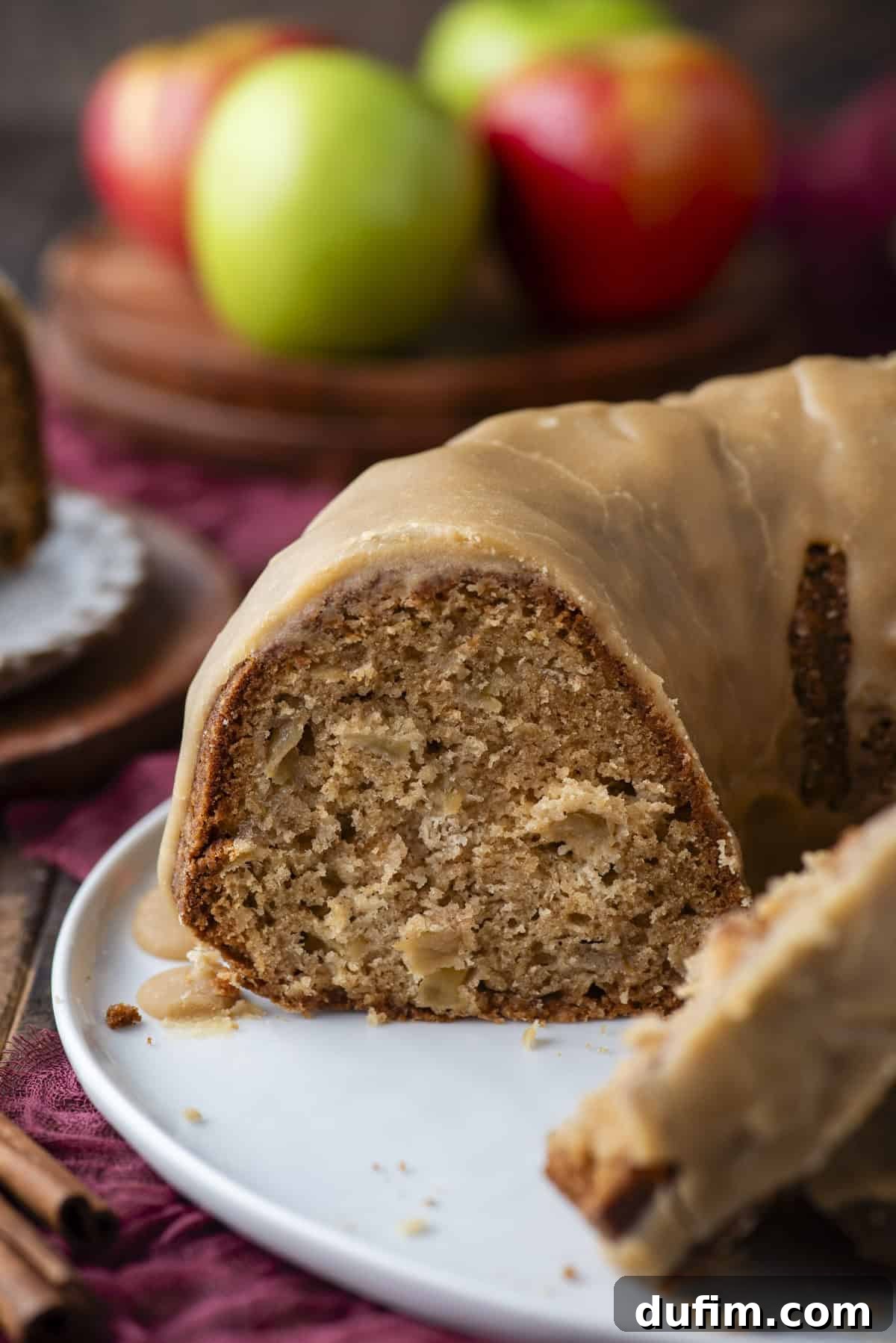 A close-up of a sliced bundt cake with caramel glaze, showing a moist, dense interior. In the background, green and red apples are slightly out of focus on a wooden plate.