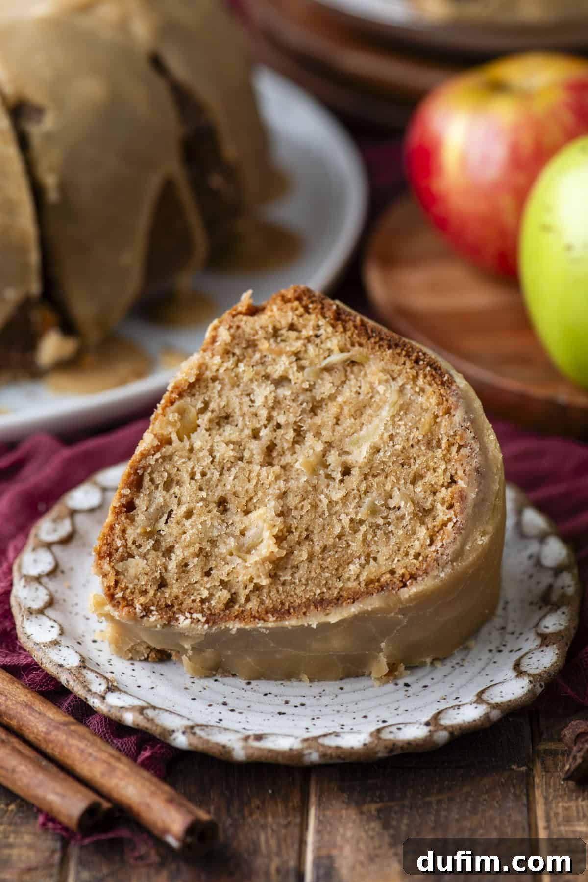 A slice of glazed apple cake sits on a ceramic plate, showing a moist, spiced interior. Whole apples and cinnamon sticks are nearby, and the rest of the cake is in the background.