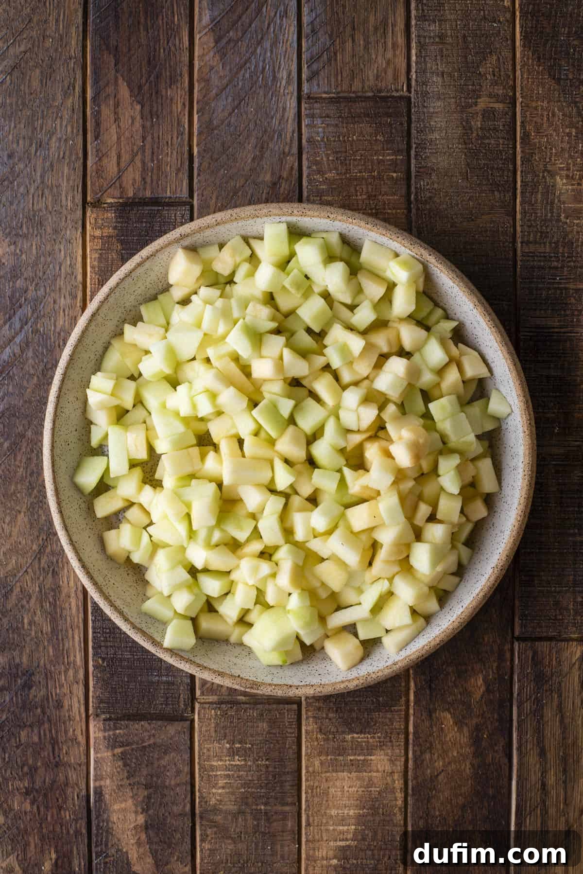 A ceramic bowl filled with small, evenly diced pieces of green apple sits on a rustic wooden surface.