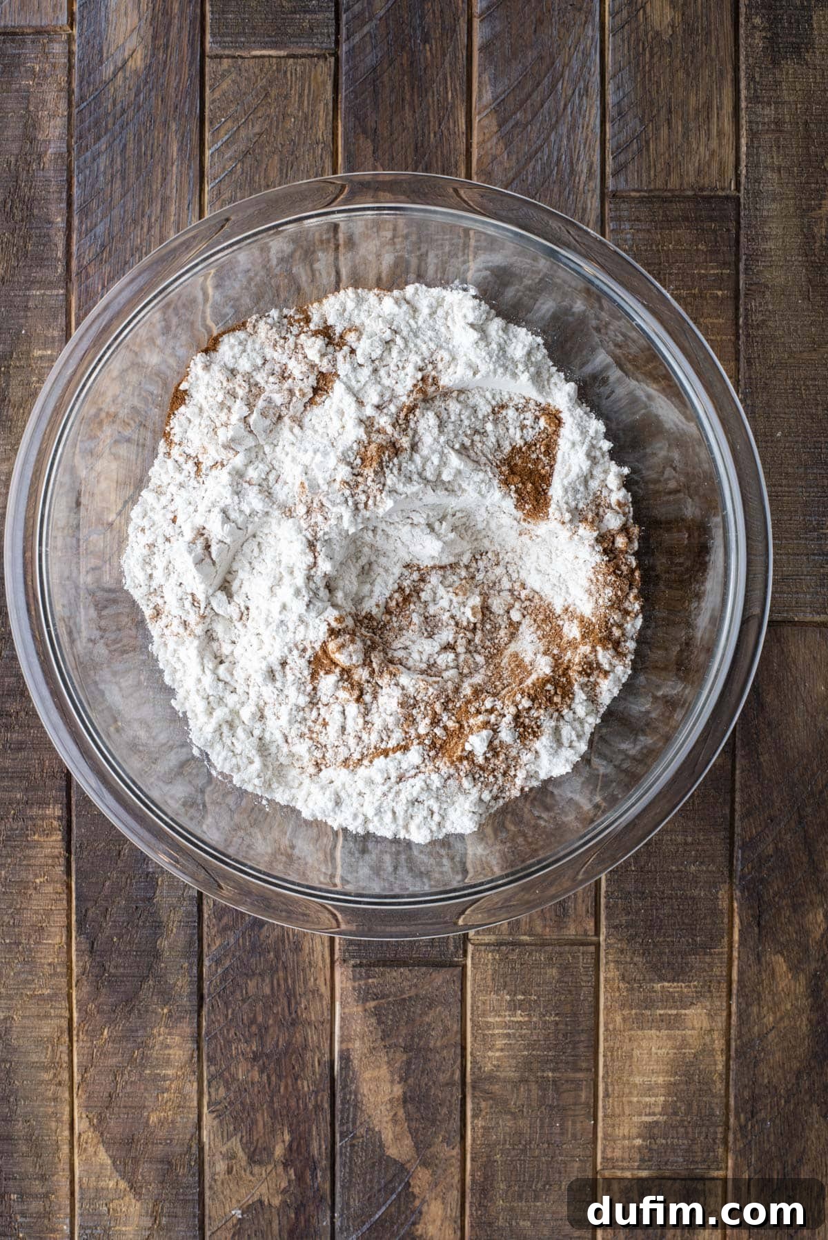 A glass bowl filled with flour and cinnamon sits on a wooden surface. The ingredients are unmixed and arranged in a mound in the center of the bowl.