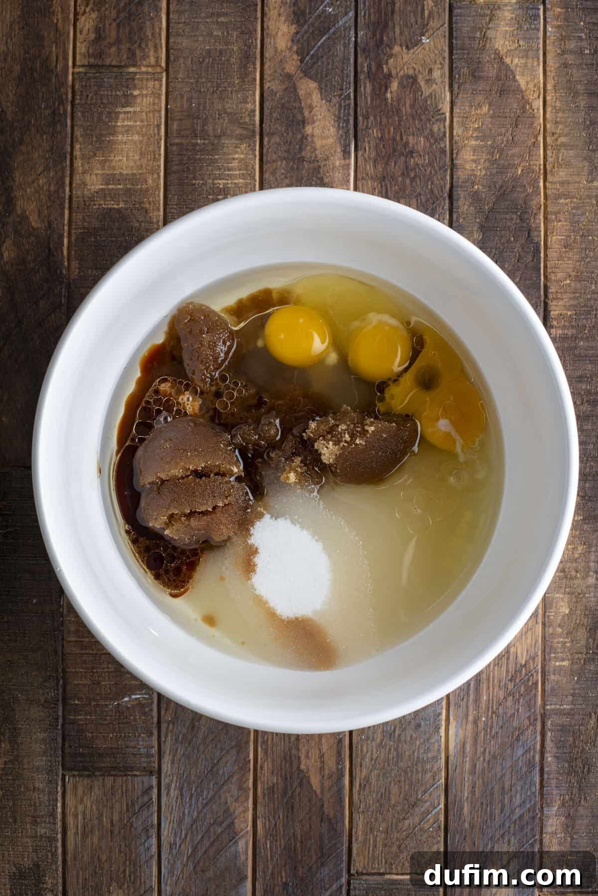 A white bowl on a wooden surface contains brown sugar, white sugar, eggs, vanilla extract, and liquid ingredients, ready to be mixed for baking.