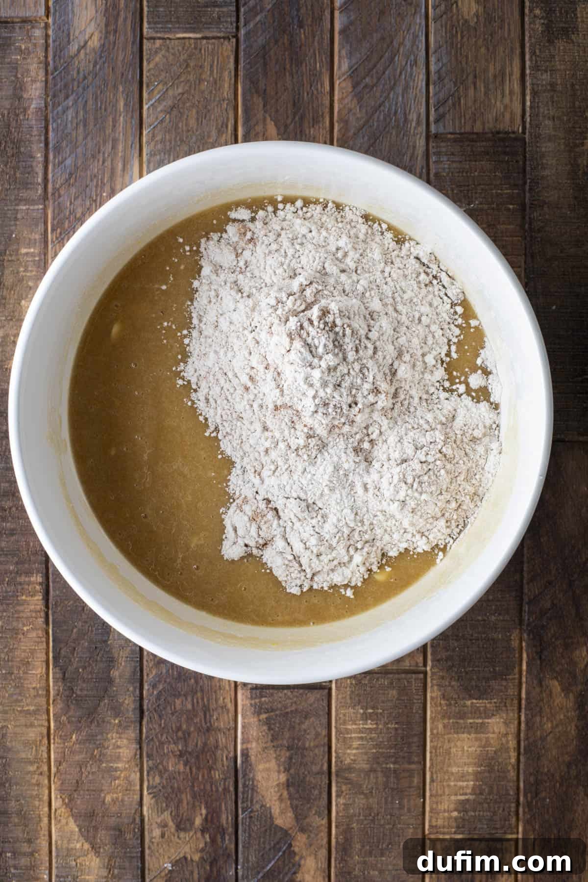 A white bowl filled with a light brown liquid mixture and topped with a mound of flour, sitting on a rustic wooden surface.