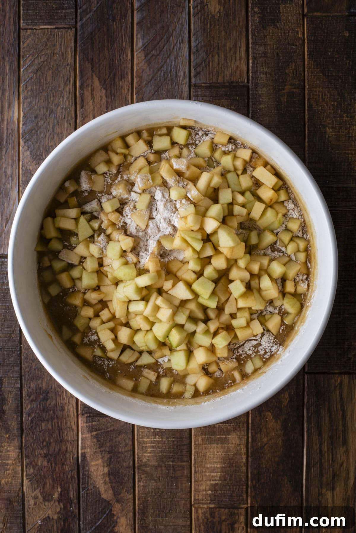 A white mixing bowl filled with diced apples, flour, and batter sits on a rustic wooden surface, ready to be mixed for baking.