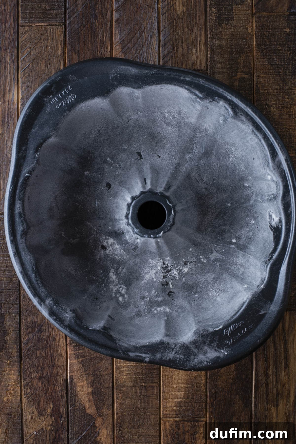 A top-down view of a flower-shaped bundt cake pan lightly dusted with flour, resting on a dark wooden surface.