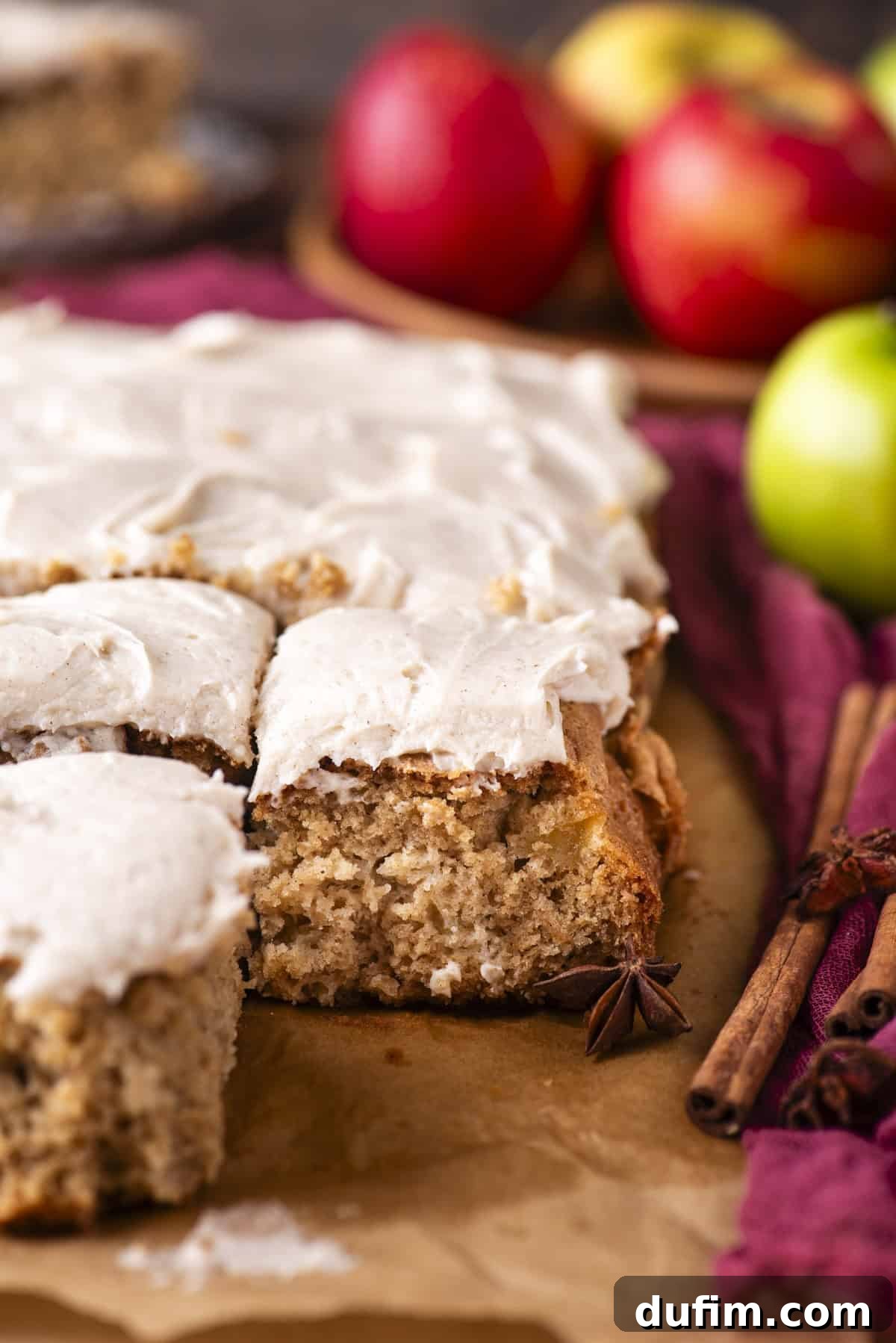 Frosted apple spice cake squares on parchment paper, with a bite taken from one piece. Cinnamon sticks, star anise, and whole apples are in the background, adding a cozy, autumn feel.