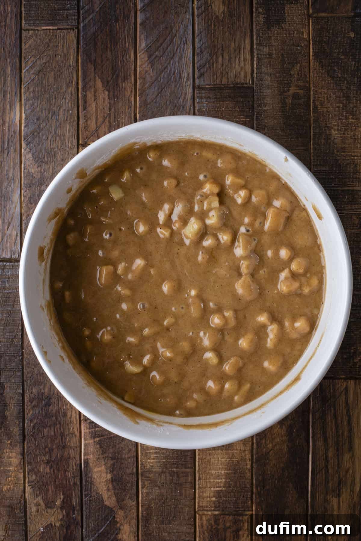 A white bowl filled with thick, chunky brown batter containing visible pieces, placed on a rustic dark wooden surface.