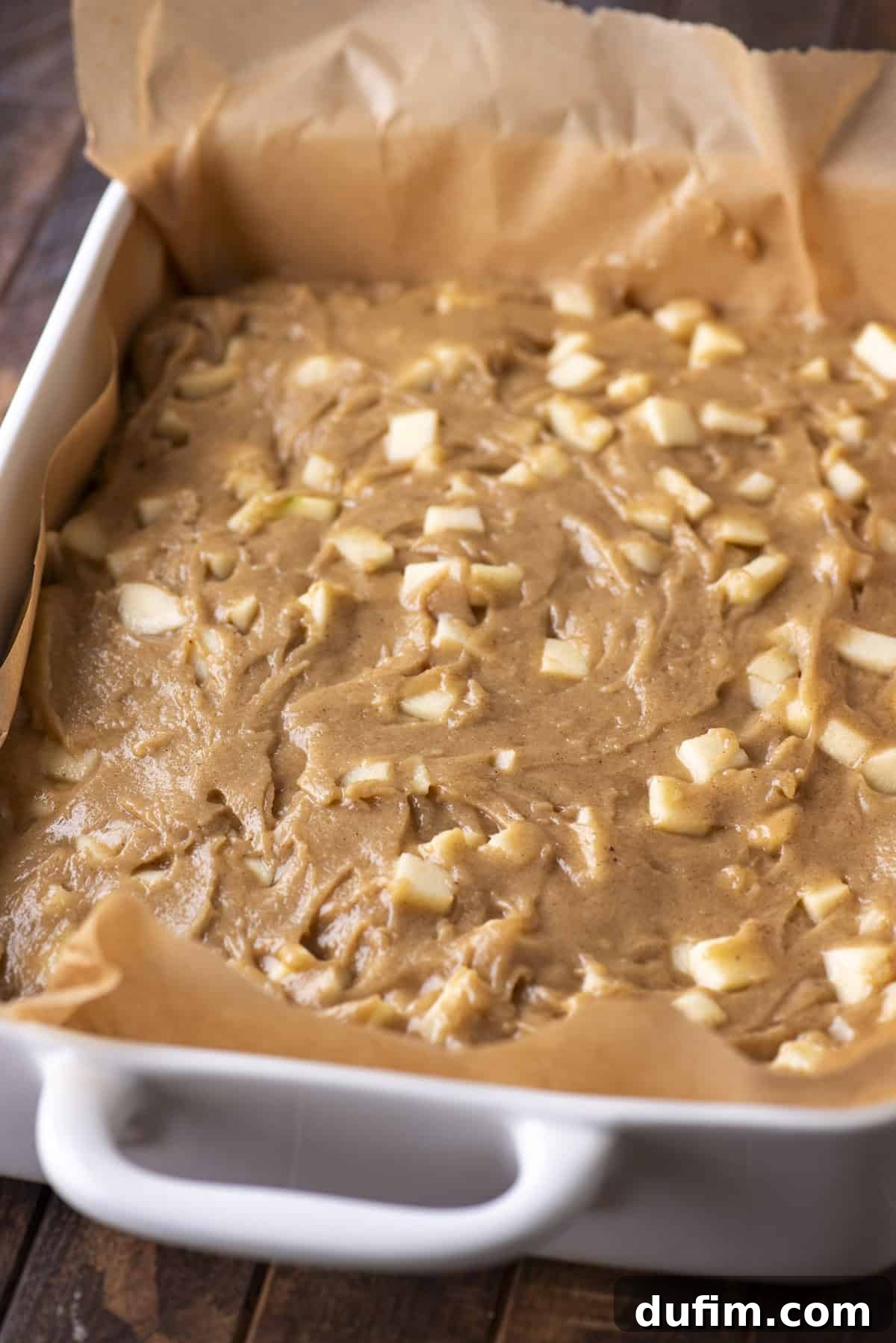 A white baking dish lined with brown parchment paper is filled with unbaked apple cake batter, showing chunks of fresh apple mixed throughout. The dish sits on a dark wooden surface.