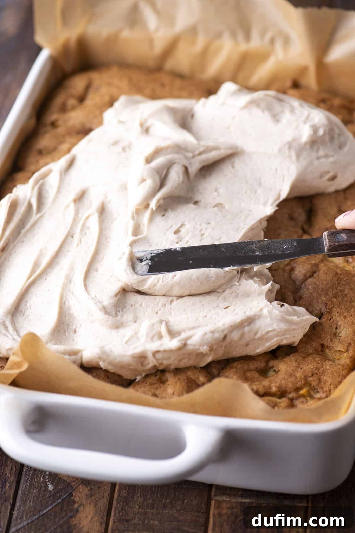 A hand spreads thick, creamy frosting over a large sheet cake in a white baking dish lined with parchment paper.