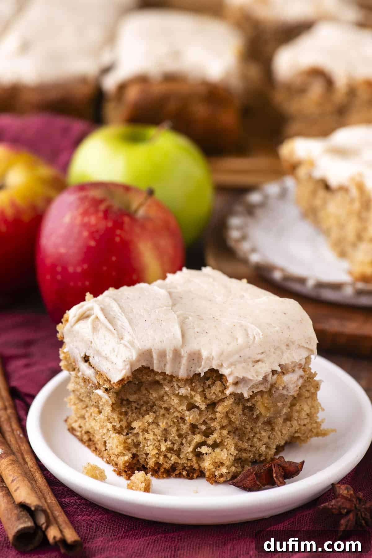 A square piece of spiced cake with creamy frosting sits on a white plate, surrounded by whole red and green apples, cinnamon sticks, and more frosted cake slices in the background.